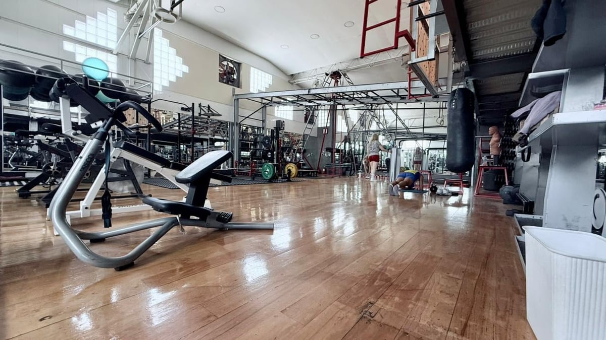 Indoor fitness facility with stationary bike in foreground, basketball hoop visible, wooden floor, and gym equipment throughout the spacious gymnasium