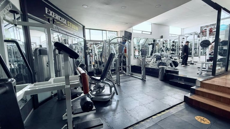 Modern gym interior with exercise equipment, machines, and mirrored walls. Several people are working out in this spacious fitness facility