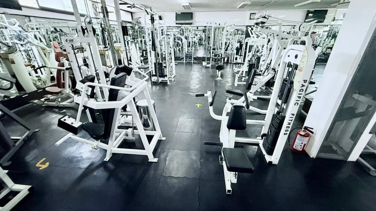 Modern gym interior with rows of white exercise machines on dark flooring, bright overhead lighting, and a spacious workout facility